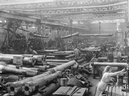 A busy howitzer shop at Coventry Ordnance Works. Men and women work together as several gun barrels are transported overhead by pulleys.