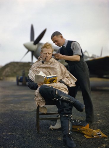 A pilot at Fairlop airfield in Essex has a haircut during a break between sweeps. A Supermarine Spitfire is in the background.
