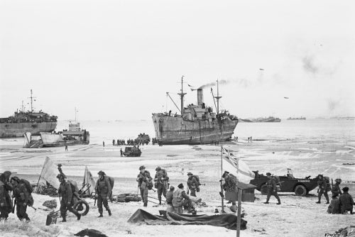 Troops come ashore on one of the Normandy invasion beaches, past the White Ensign of a naval beach party, 7 June 1944.