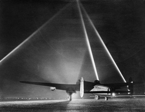 An Avro Lancaster Mk III of No. 103 Squadron pauses on the flarepath at Elsham Wolds, Lincolnshire, before taking off for a raid on Duisburg, 26 March 1943. In the background three searchlights form a cone to indicate the height of the cloud base.